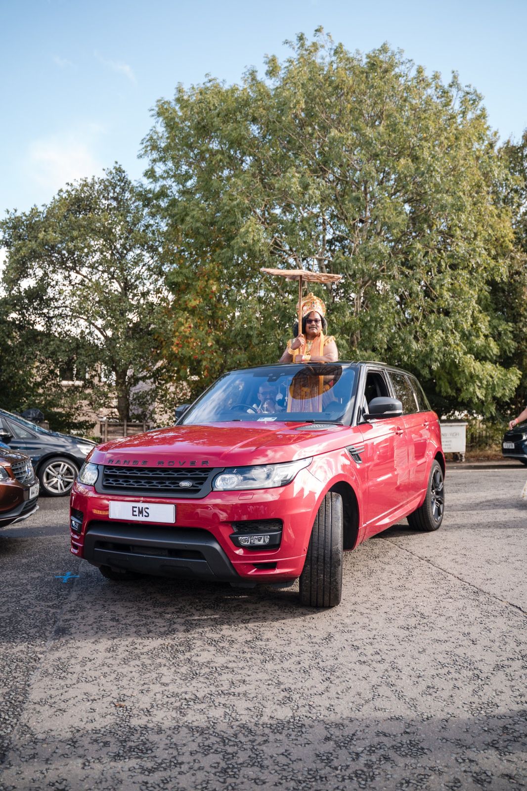 Maveli in golden crown with umbrella standing through the sunroof of a red Range Rover