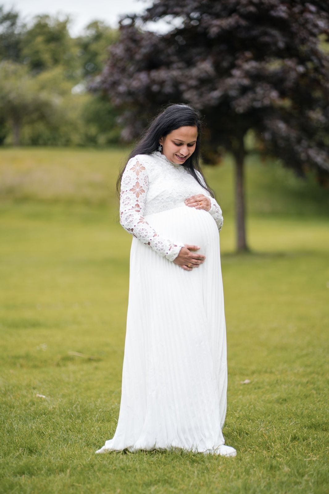 Mother-to-be smiling down at her bump beside a copper beech tree